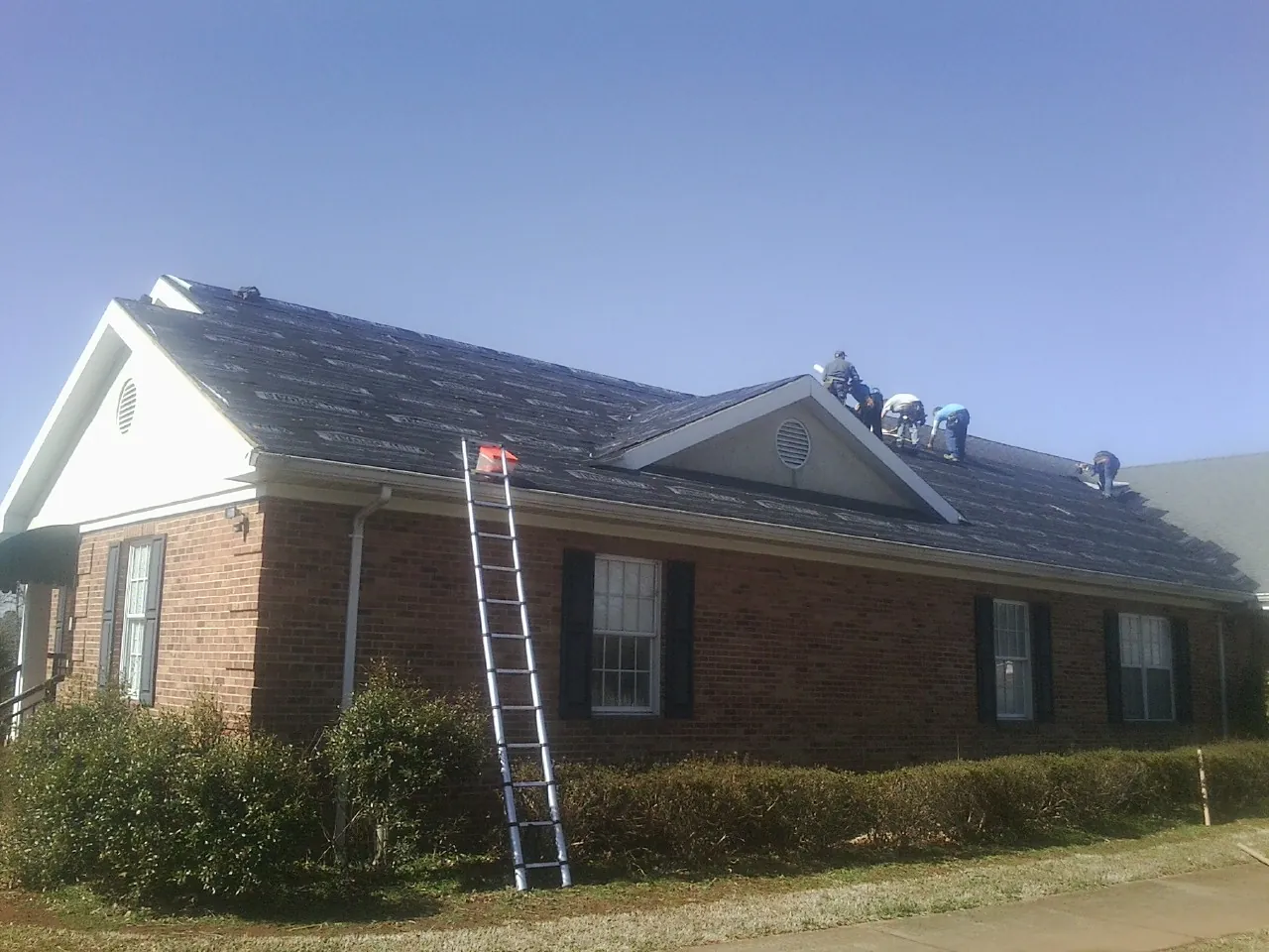 Crew tearing shingles off a brick ranch roof with a ladder set — Emergency Roof Repair work by our crew in Mc Clelland