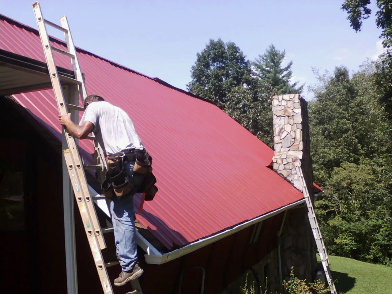 Roofer on a ladder beside a red metal roof and stone chimney — Soffit & Fascia Repair by Roofing Brothers in Mc Clelland